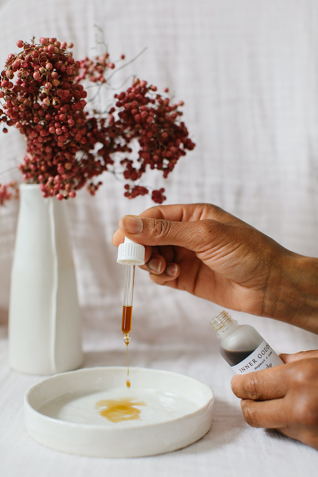 Forage Botanicals Inner Goddess Tincture. Vegan hormone support to help reset your monthly cycle and regulate irregular periods A person is holding the bottle in one hand and using the pipette to drop the tincture on a small white ceramic plate. There are dried red flowers in a white ceramic vase behind.