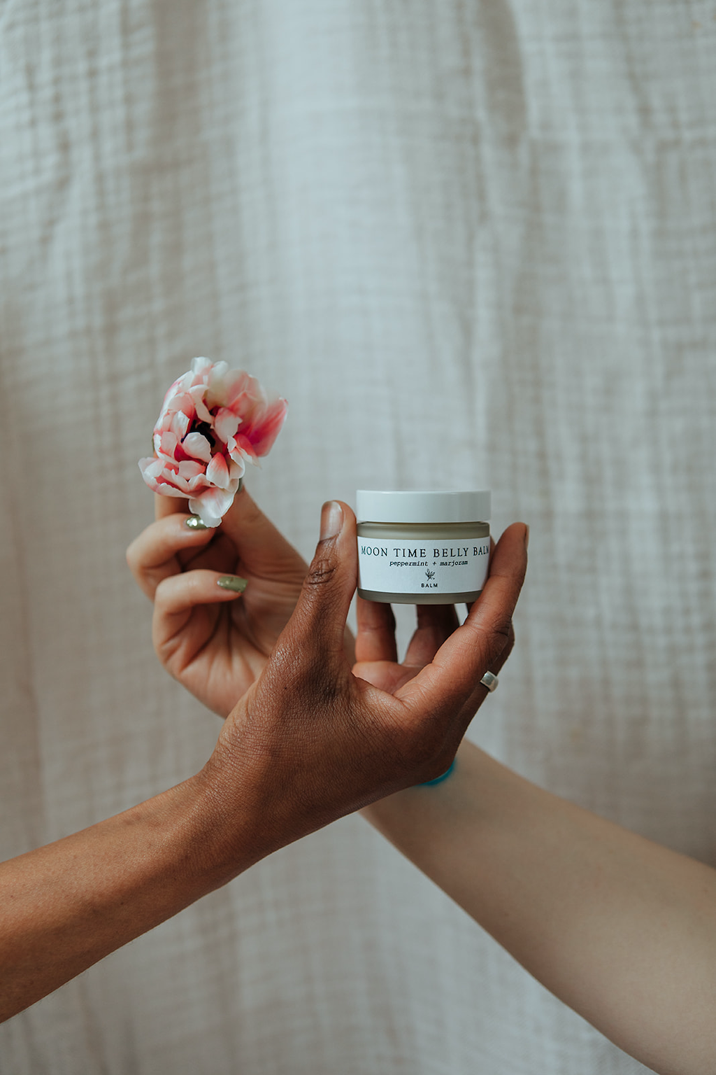 Forage Botanicals Moon Time Belly Balm. Vegan menstrual cramp relief. One hand is holding the jar of balm, while another hand crosses it from behind holding a pink flower