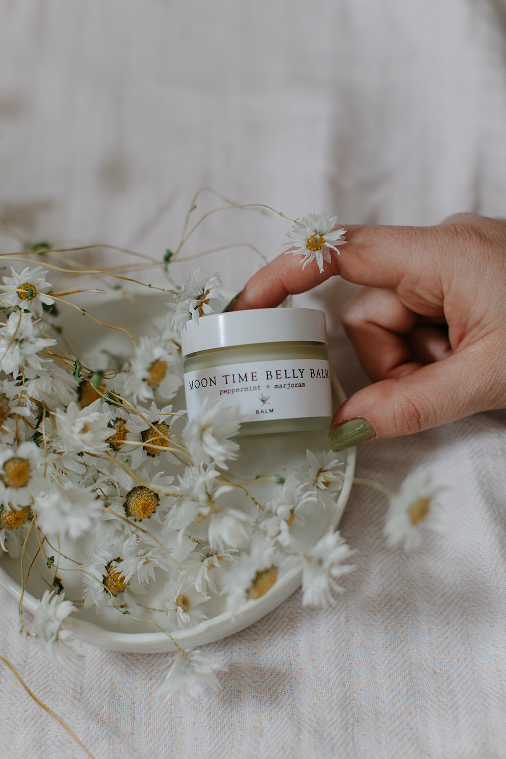 Forage Botanicals Moon Time Belly Balm. Natural menstrual pain relief. A person is tilting the jar up label forward on a small white ceramic plate next to a bunch of daisies.