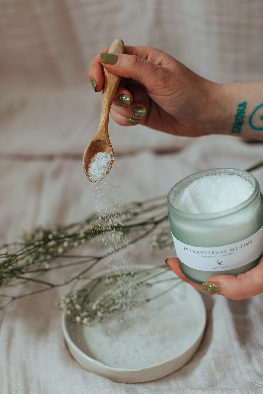 Forage Botanicals Premenstrual Me Time Bath Salts. Vegan menstrual cramp relief. A person is holding the jar in one hand and gently spilling a wooden spoon of salts onto a white ceramic plate below with some sprigs of dried flowers nearby.