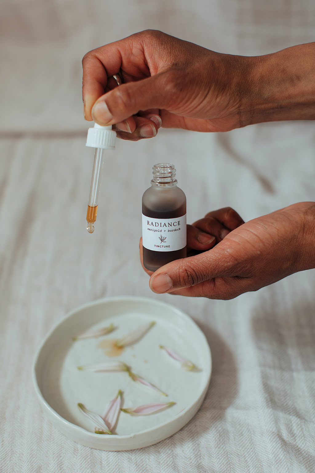 Forage Botanicals Radiance Tincture. Vegan hormone help. A person is holding the bottle in one hand and using the pipette to drop the tincture on a small white ceramic plate. There are dried flower petals on the plate.