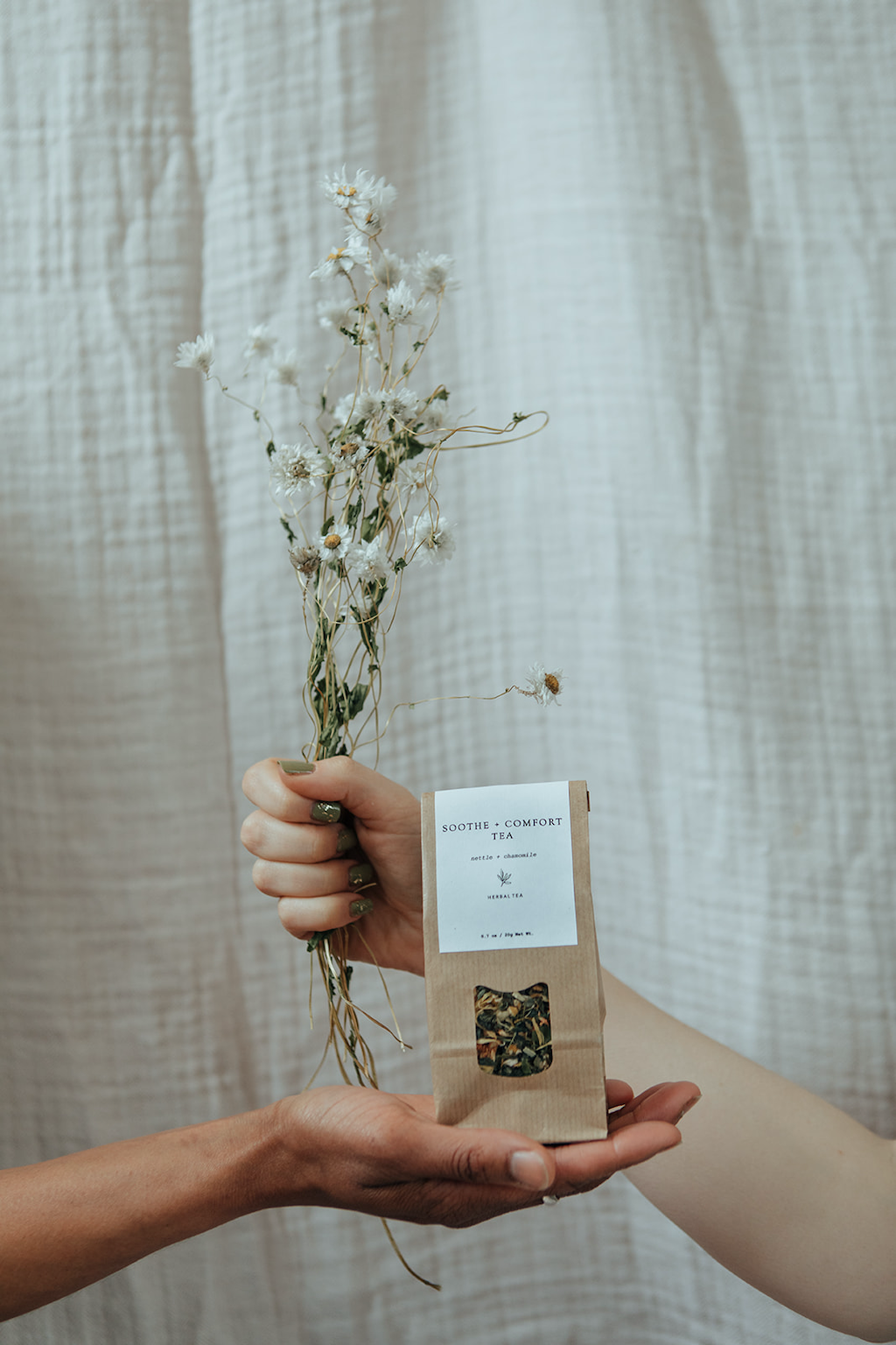 Forage Botanicals Soothe and Comfort Tea. Vegan stress relief tea. One hand is holding the bag of tea in their palm, while another hand crosses it from behind holding a bunch of dried chamomile flowers.