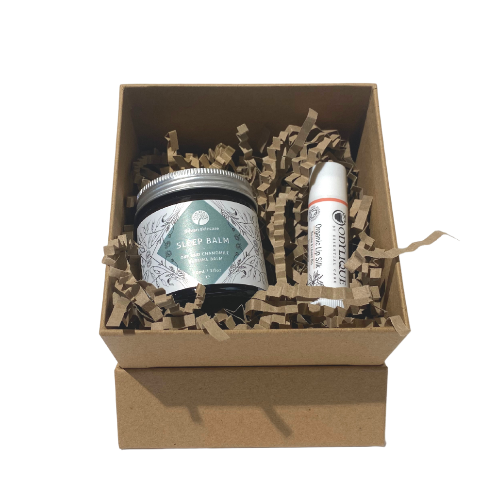 brown kraft cardboard gift box sitting propped on its lid lid in a small cube shape filled with brown kraft paper shred. inside the box there is a sleep balm and a lip balm stick