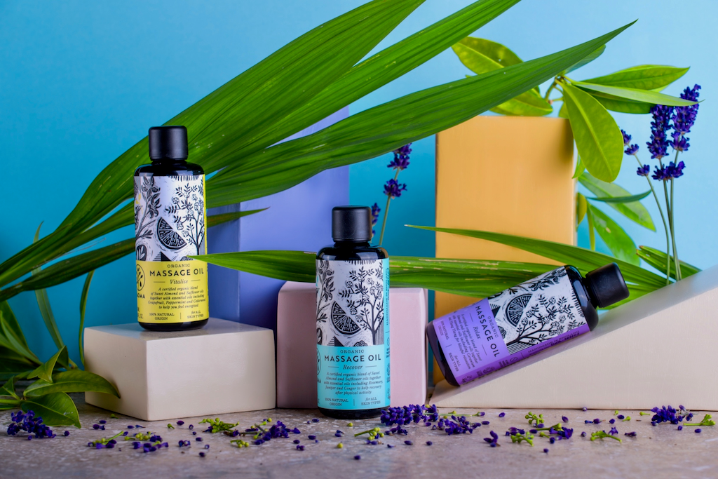 Haoma Organic Massage Oils. Cruelty free body oils. The massage oils are packaged in a black glass bottles with coloured labels and black illustrations of plants. They are displayed on a variety of coloured plinths with various plant leaves and sprigs of bright purple lavender strewn around.