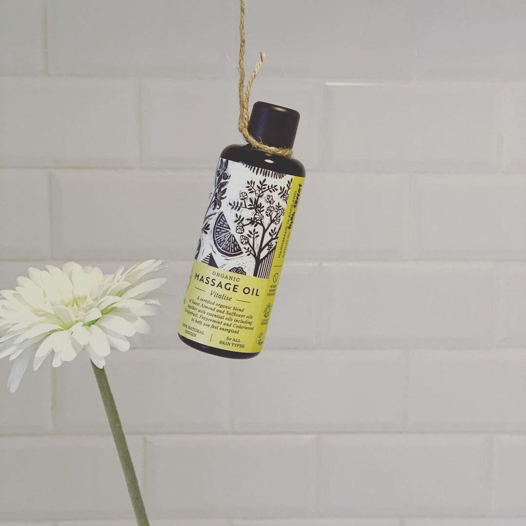Haoma Organic Massage Oil in Vitalise. Natural body oil for energy. The massage oil is packaged in a black glass bottle with a yellow label and black illustrations of plants. The bottle is hanging from a piece of twine in front of a white ceramic tile wall with a white flower next to it.