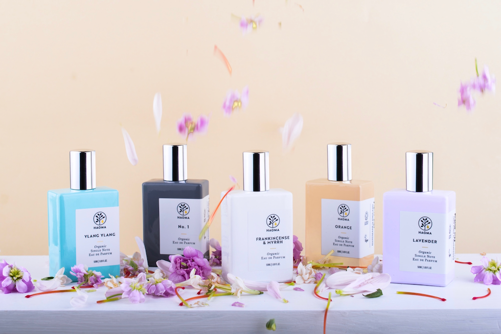 Haoma Organic Eau de Parfum. Certified organic perfumes. The perfume bottles are pictured on a white shelf with flowers and flower petals all around them.
