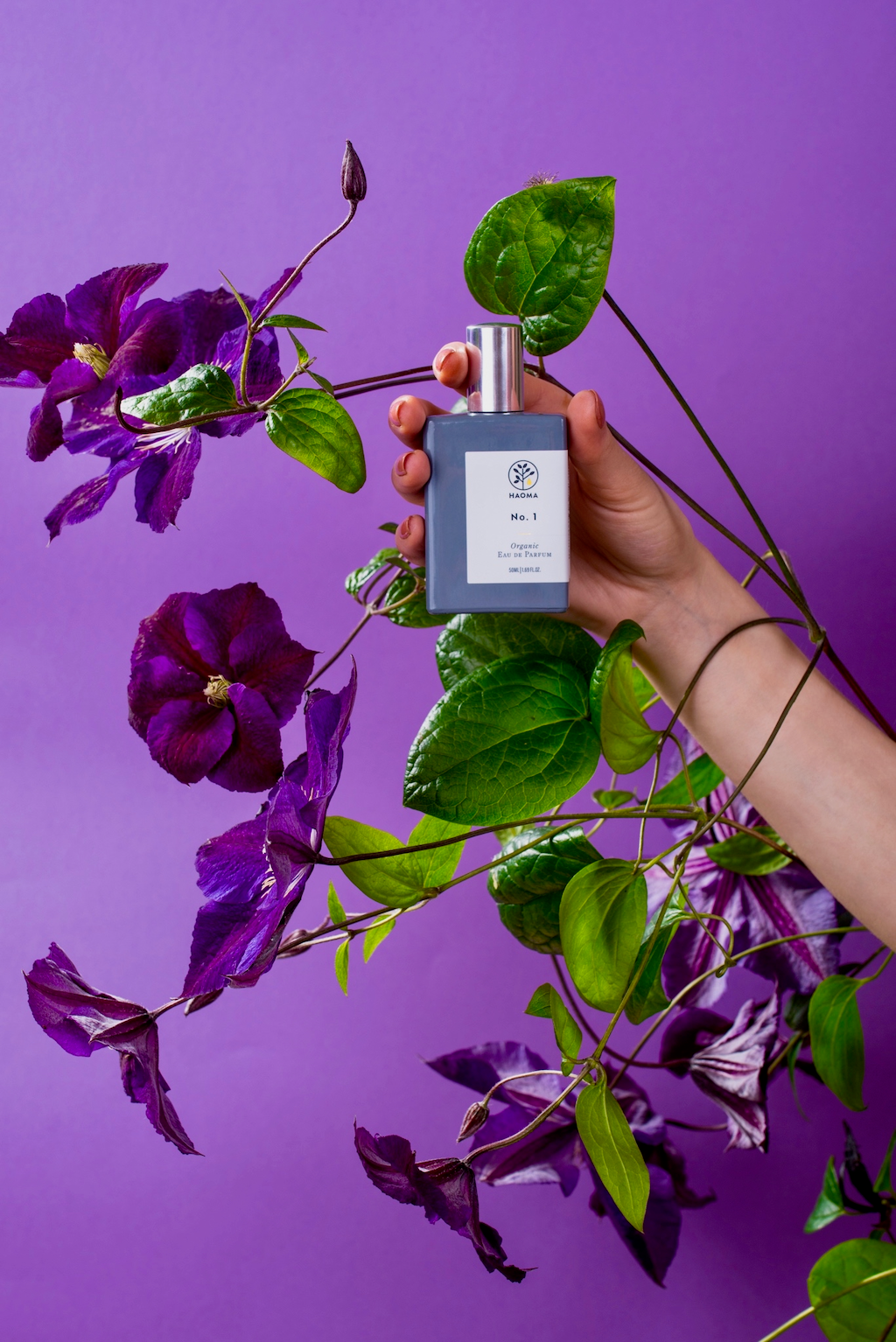 Haoma Organic Eau de Parfum. Certified vegan perfumes. The No.1 Blend bottle is shown held in someone's had with a vine of leaves and purple flowers wrapped around and behind.