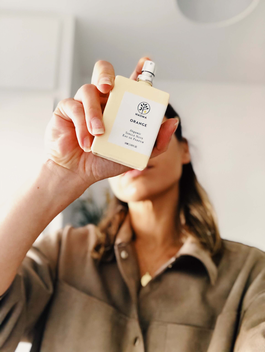 Haoma Organic Eau de Parfum. Certified organic perfumes. The Orange bottle is shown being held in someone's hand in the foreground as if they are about to spray it forward, with the hand and bottle covering their face in the background.