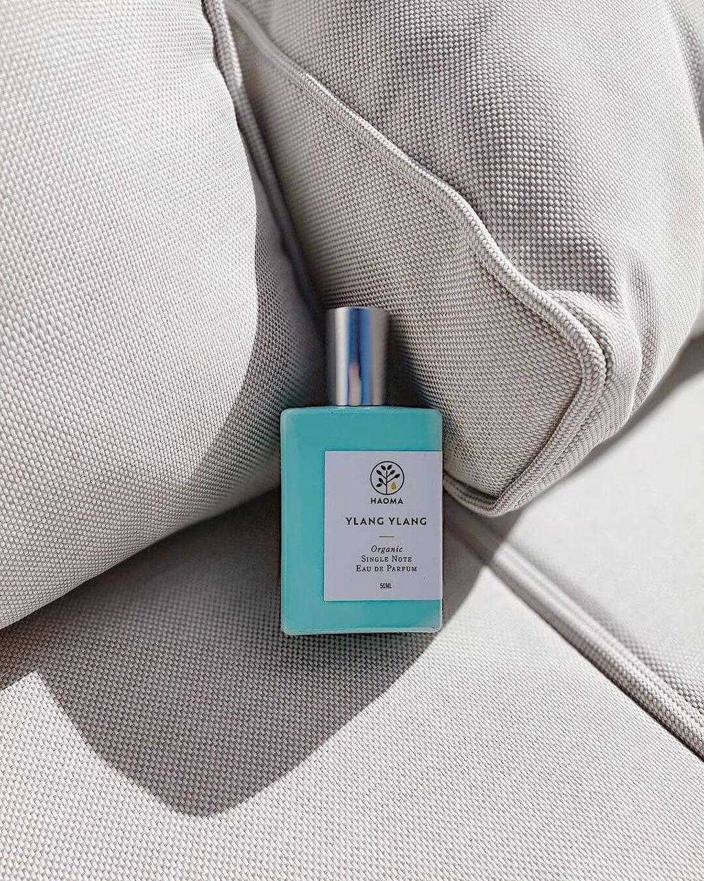 Haoma Organic Eau de Parfum. Certified organic eau de parfum. The Ylang Ylang bottle is shown on some grey sofa cushions in the sun.