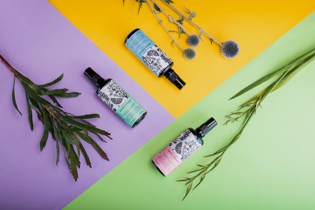 Haoma Organic Spray Deodorant. Natural deodorant without bicarbonate of soda. The deodorants are sitting against their colour wheel opposing backgrounds with plants next to them.