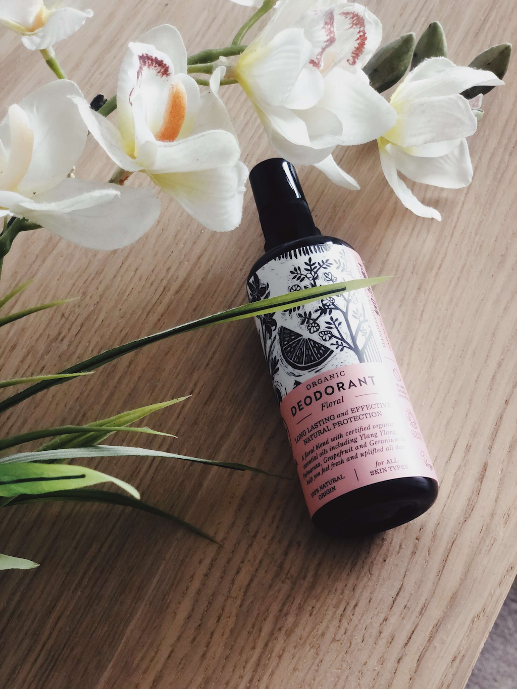 Haoma Organic Spray Deodorant. Certified vegan deodorant without bicarbonate of soda. The Floral deodorant is sitting on a wood table with white flowers draped over it.