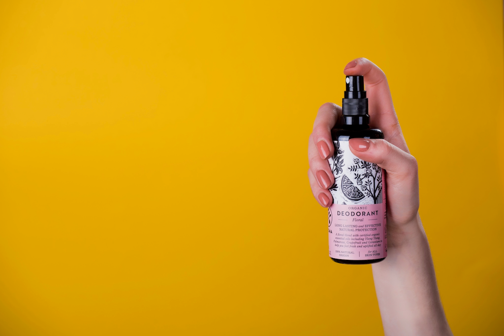 Haoma Organic Spray Deodorant. Natural deodorant without bicarbonate of soda. A hand with pink painted nails is holding the Floral deodorant ready to spray against a yellow background.