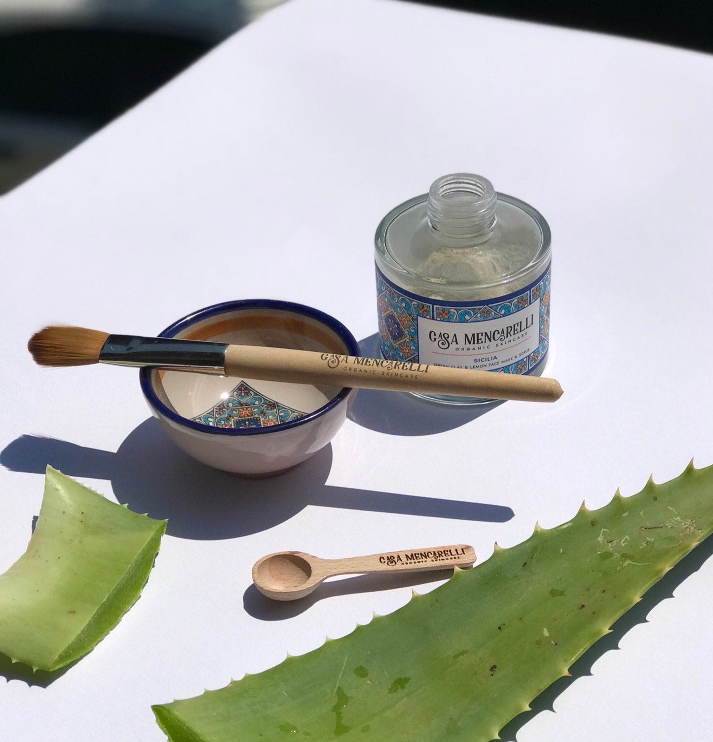 Pictured is the casa mencarelli mask mixing bowl with the mask mixing brush resting on top of the bowl. Placed diagonally behind in the green clay and lemon mask in its glass bottle. In the foreground is an Aloe Vera leaf and the casa mencarelli wooden spoon