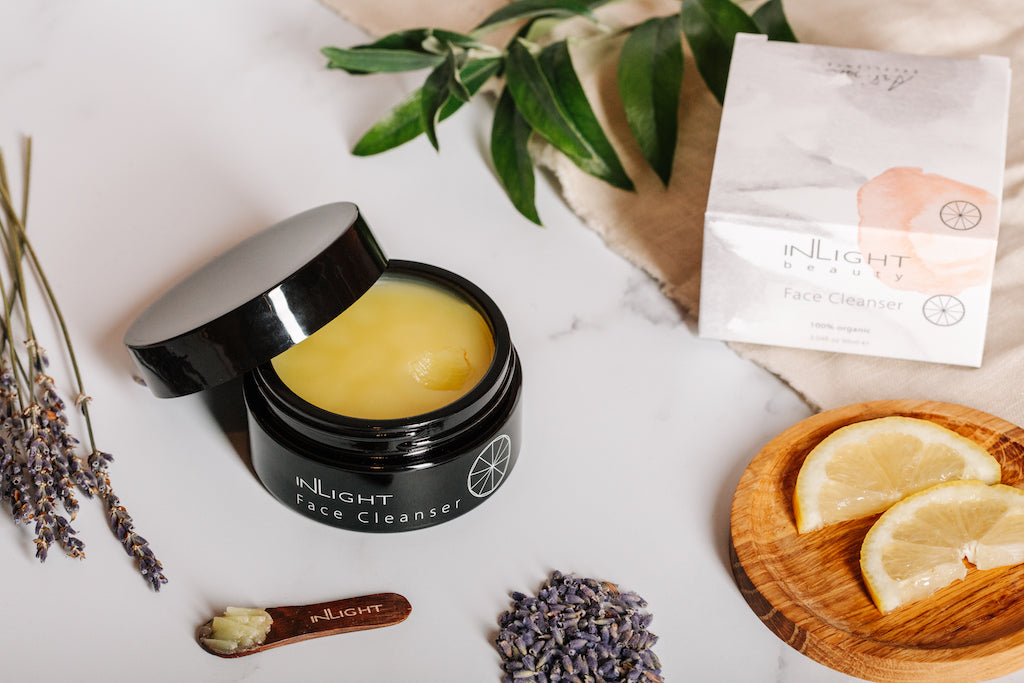 flatlay of inlight beauty's face cleanser balm. the open pot of cleanser can be seen surrounded by some fresh ingredients that are in the product. 2 slices of lemon on a wooden coaster, some dried lavender in a pile and also on the stem and a sprig of bay leaves. there is an organic fabric laying under the leaves which the box for the cleanser is sitting on