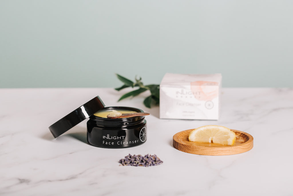 lifestyle image of inlight beauty 100% organic cleansing balm which can be seen sitting on a white and grey marbled surface. the black glass jar is open with the black plastic lid resting on top of it alongside a wooden spatula which has some of the creamy yellow coloured cleansing balm on it. in the foreground there is some dried lavender. to the right of the jar is a wooden coaster with a slice of lemon on it. in the background you can see the box for the cleanser blurred sitting next to some greenery