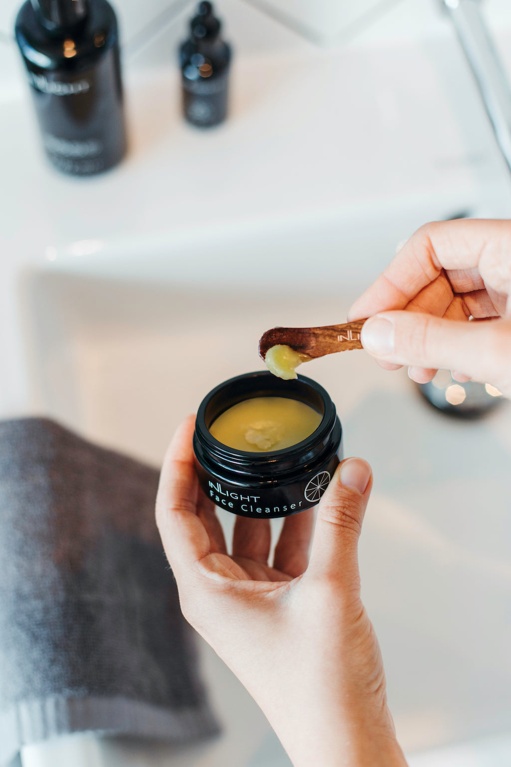 lifestyle photo of inlight beauty's 100% organic cleansing balm. the cleansing balm is being held in someone's hands. in the left hand is the open black glass jar and you can see the golden coloured balm inside. in the right hand is a wooden spatula with some cleansing balm on it. the jar is being held over a sink and you can see blurred in the background a grey towel draped over the white sink and some other black glass bottles of inlight beauty products