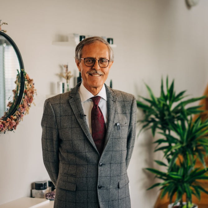 dr spiezia founder of inlight beauty who is standing in a grey checked suit in a hallway with white paint, a black mirror surrounded by pink flowers and a spiky green palm plant and pine coloured stairs in the background