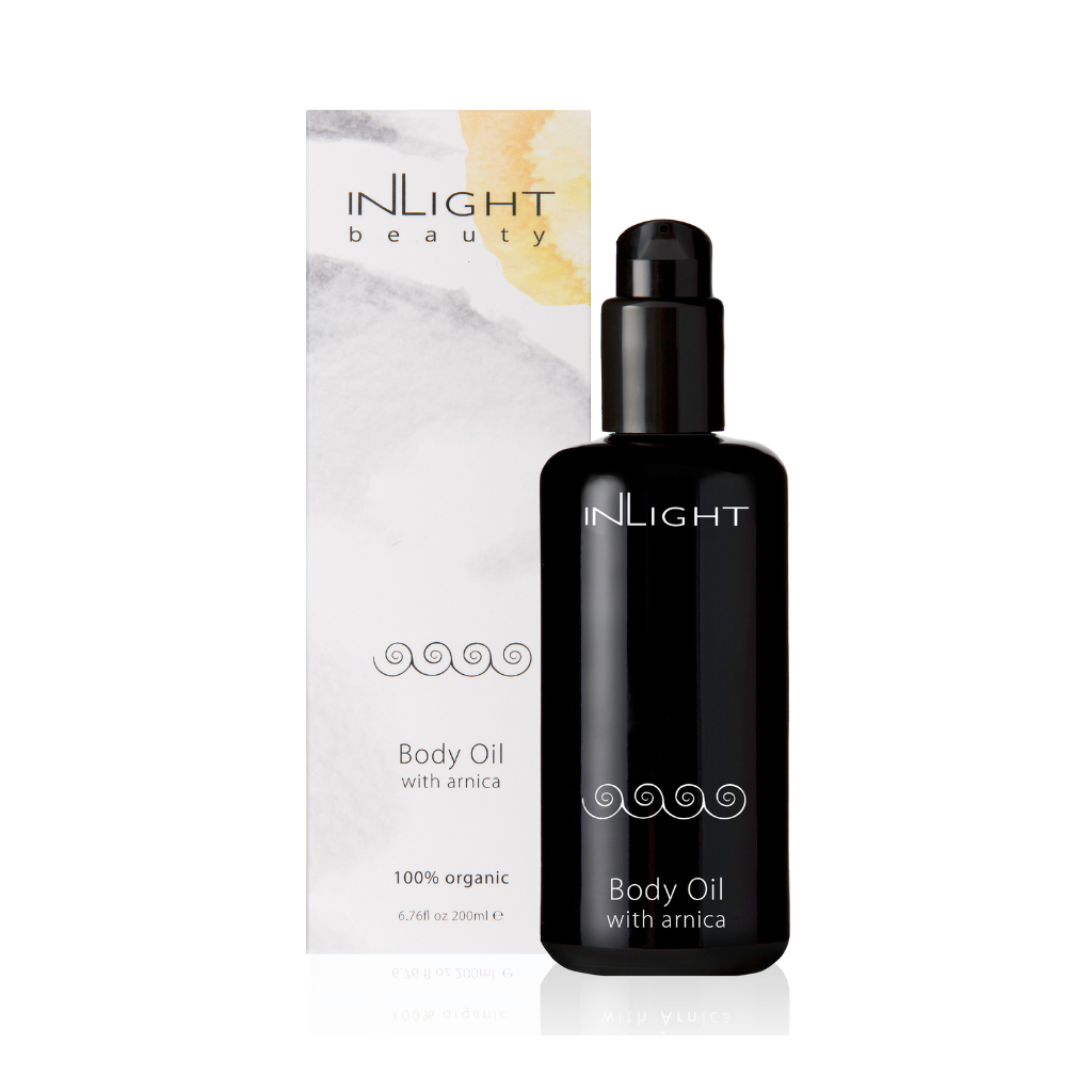 inlight beauty's body oil with arnica is displayed on a white background there is a tall box that is predominantly white with soft splashes of grey and yellow. the body oil itself is next to the box and is in a black bottle with a black pump