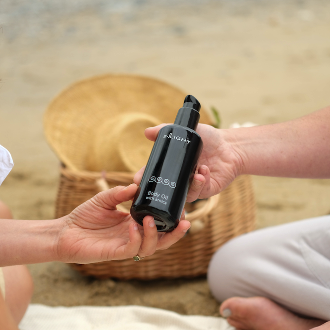 body oil containing arnica is being passed from one hand to another by two people sitting cross legged on a beach. in the background there is a white picnic blanket and a wicker basked with a natural fibre sun hat placed on top
