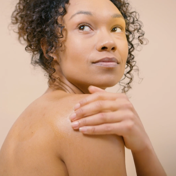 woman with darker toned skin and dark brown corkscrew curly hair massaging body oil into her shoulder looking upwards