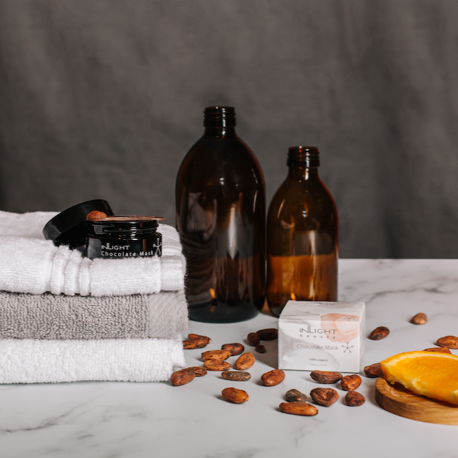 inlight beauty's 100% organic chocolate face mask sat on a bed of white and grey fluffy towels next to empty amber glass bottles. The pink, grey and white box for the mask is to the right of the towels and surrounded by cocoa pods and an orange slice which are both ingredients in the face mask