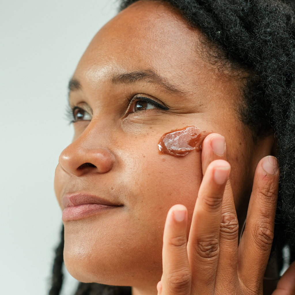 woman with brown skin and black dreadlocked hair applying inlight beauty's 100% organic chocolate face mask to her cheek with her fingers in side profile