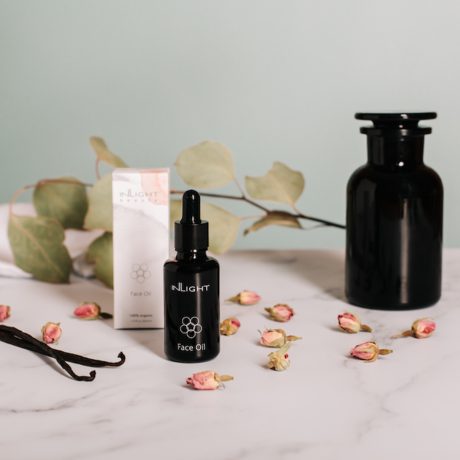 inlight beauty 100% organic face oil can be seen on a grey and white marbled surface amongst dried rosebuds, some greenery, vanilla pods and next to a black glass apothecary jar