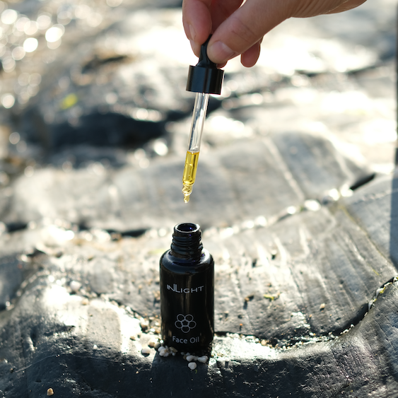 inlight beauty facial oil sitting on some black rocks on the beach with some smaller pebbles sitting in the grooves of the rock. the black glass bottle is open and someone is holding the pipette above the bottle as if they are about to dispense the face oil to be used
