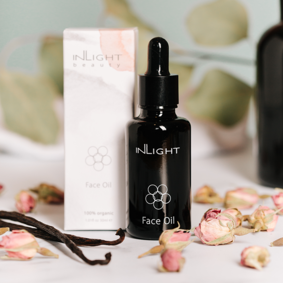close up of inlight beauty face oil in black gloss pipette bottle. in the background there are some green leaves and another black glass bottle. in the foreground are dried rose buds and vanilla pods