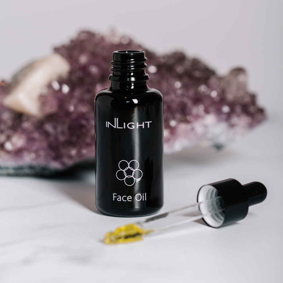 inlight beauty 100% organic facial oil can be seen in its black glass bottle with the pipette sitting in the foreground with some of the golden colour oil in the dropper. in the background is an amethyst crystal