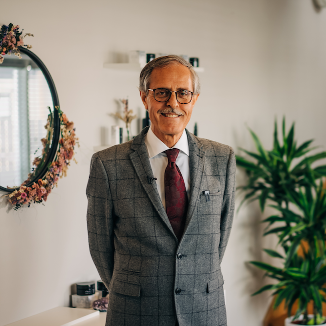 dr spiezia founder of inlight beauty who is standing in a grey checked suit in a hallway with white paint, a black mirror surrounded by pink flowers and a spiky green palm plant and pine coloured stairs in the background