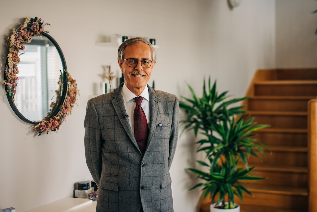 dr spiezia founder of inlight beauty who is standing in a grey checked suit in a hallway with white paint, a black mirror surrounded by pink flowers and a spiky green palm plant and pine coloured stairs in the background