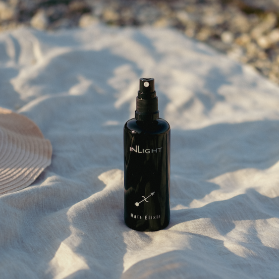 black bottle of inlight beauty hair oil on a grey linen blanket with a wicker hat and a pebbled beach in the background