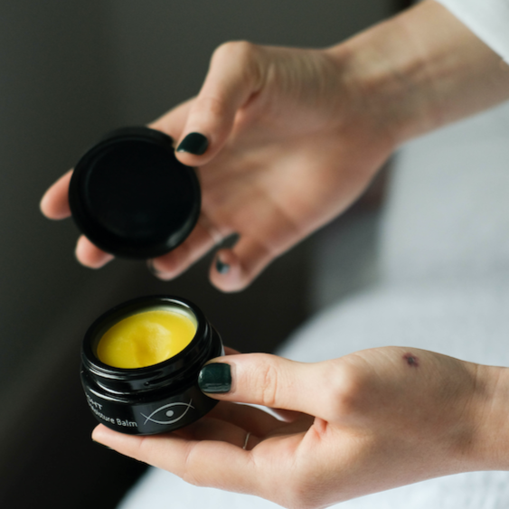 inlight beauty's deep moisture balm is held in a woman's hands who has green nail varnish on. She is holding the jar which is open in one hand and the lid in the other as if she has just opened it. You can see the yellow texture of the balm inside the jar