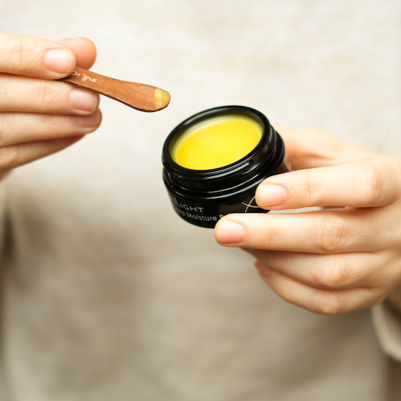 inlight beauty deep moisture balm in a black jar which is open so you can see the yellow colour and smooth texture inside. the jar is being held in someone's fingertips and they are holding a wooden spatula in their other hand which has some of the balm on it. They have light grey or fawn coloured jumper in the blurred background