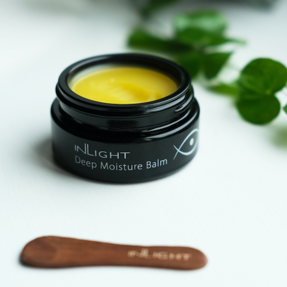 Black glass jar without a lid on so you can see the yellow buttery balm texture of inlight beauty's deep moisturising balm. in the foreground there is a small wooden spatula and some trailing green plant fronds