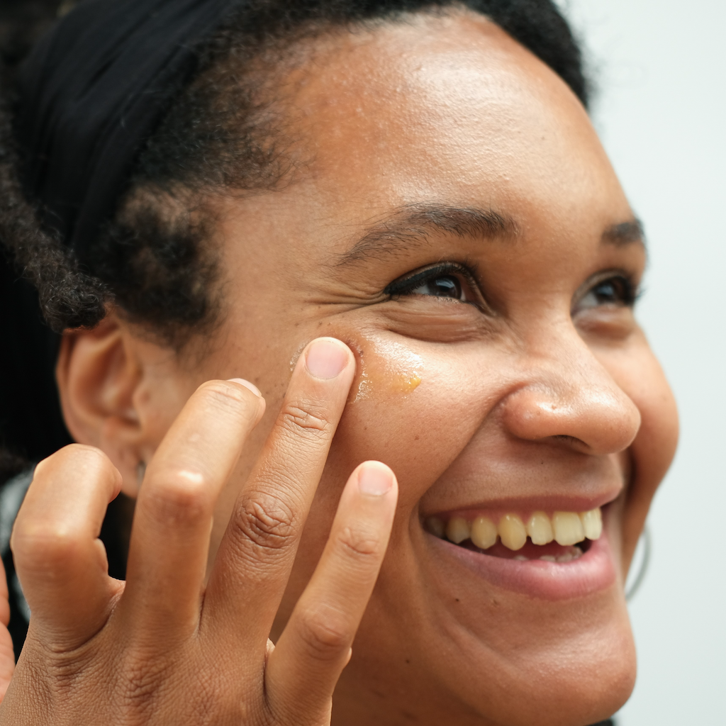 lady with dark skin tone and black textured hair smiling as she applies inlight beauty's deep moisture balm on to her cheeks with her fingertips