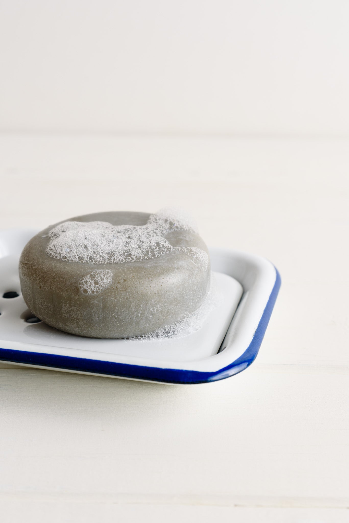 The round grey shampoo bar, foamy and bubbly, placed on an enamel tray, against on a white background