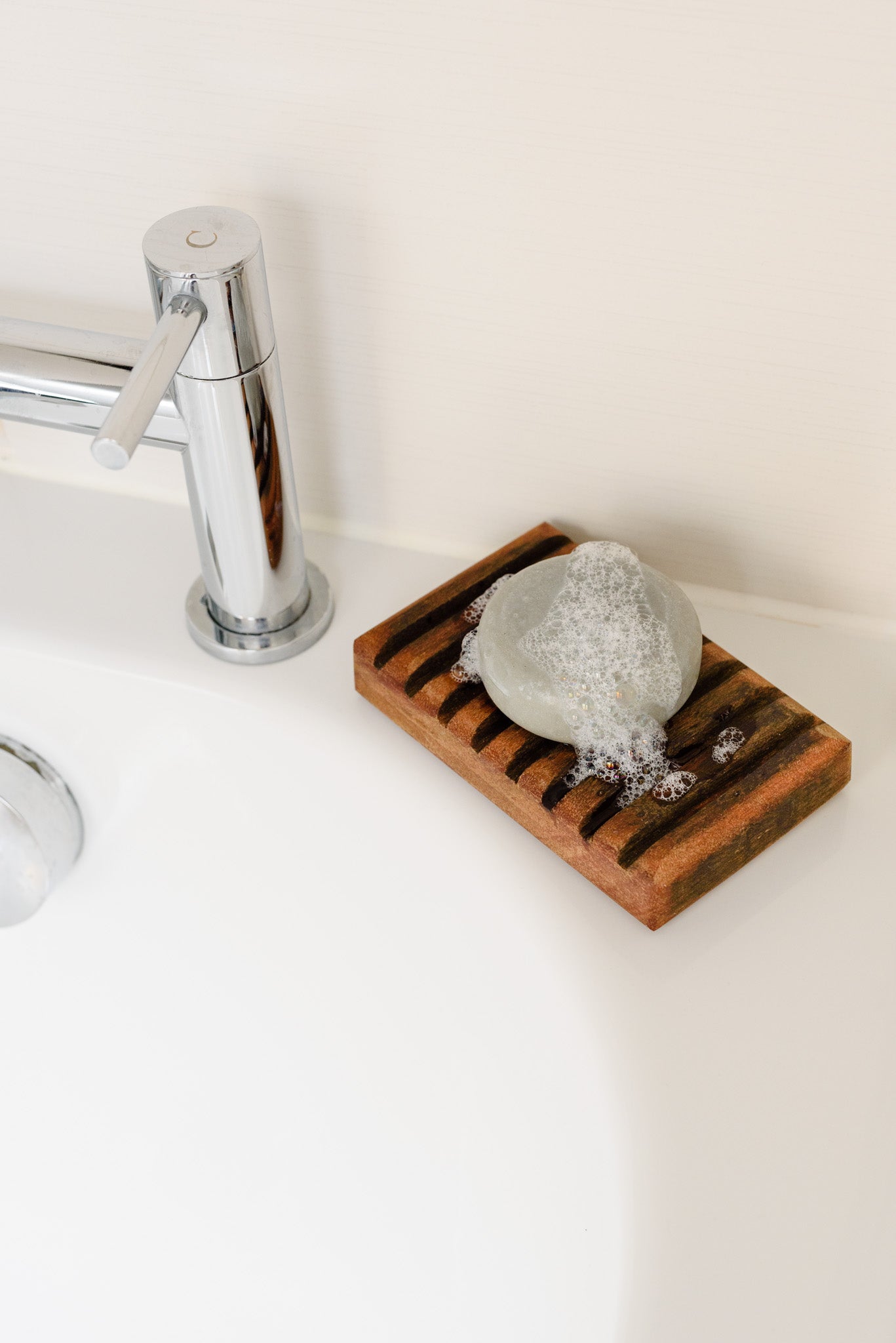 The round grey shampoo bar, foamy and bubbly, placed on a wooden tray, resting on a white sink, next to a silver tap.