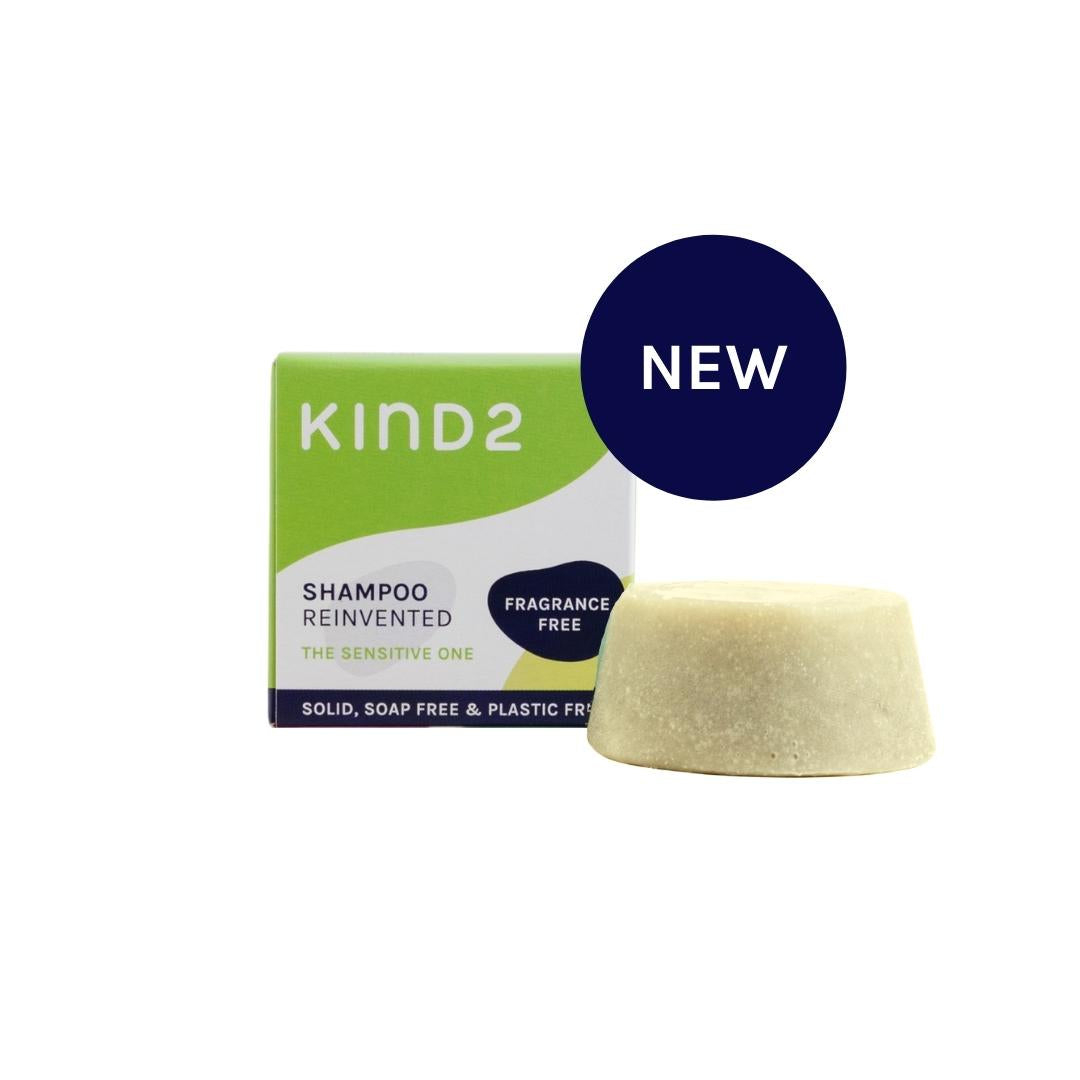 A small round off white plastic free KIND2 shampoo bar. Placed in front of its small green, white and navy box. A NEW Sticker on top of them. 