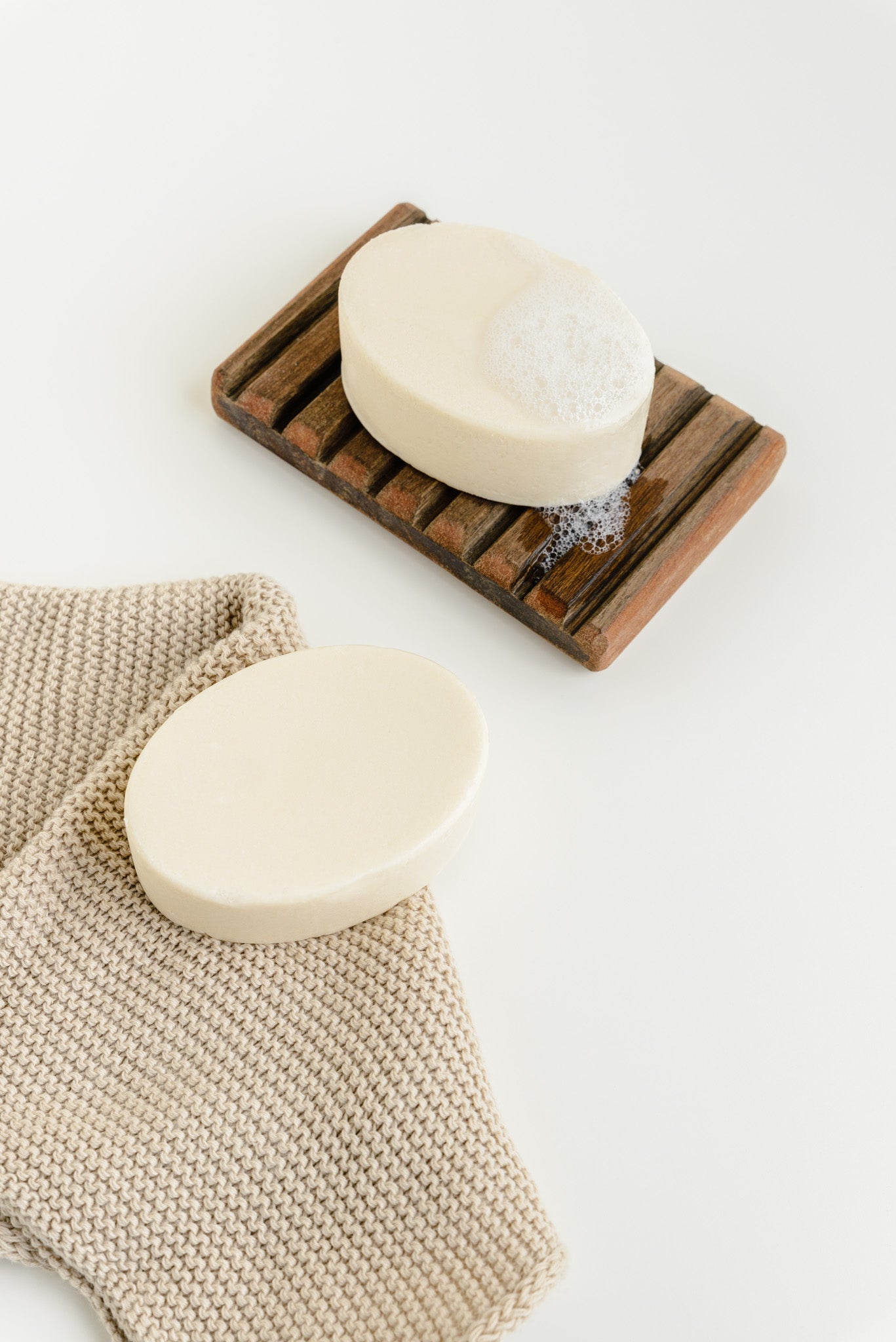 2 shampoo bars pictured against a white background. 1 is wet with foam and placed on a wooden soap tray. The other is placed on a beige cloth.