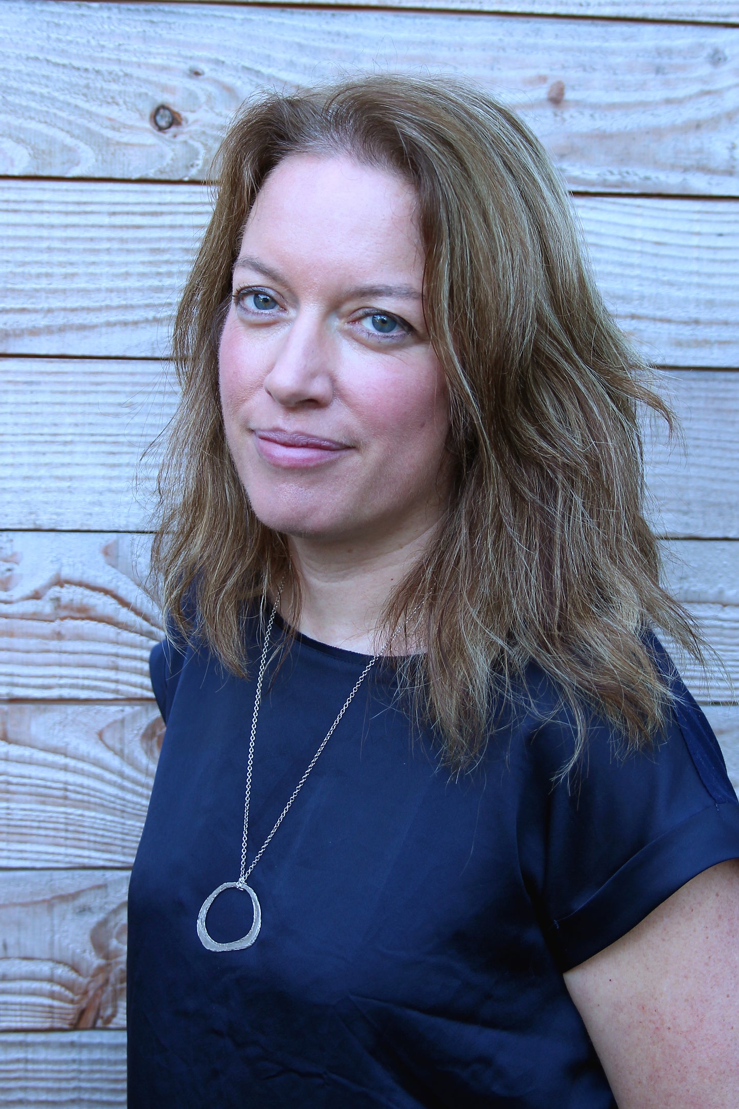 photo of marian who makes silvan skincare vegan face creams from her studio in the welsh borders. Photo of marian standing in front of natural wood panelling with light brown hair and a blue boat neck top with a gold chain