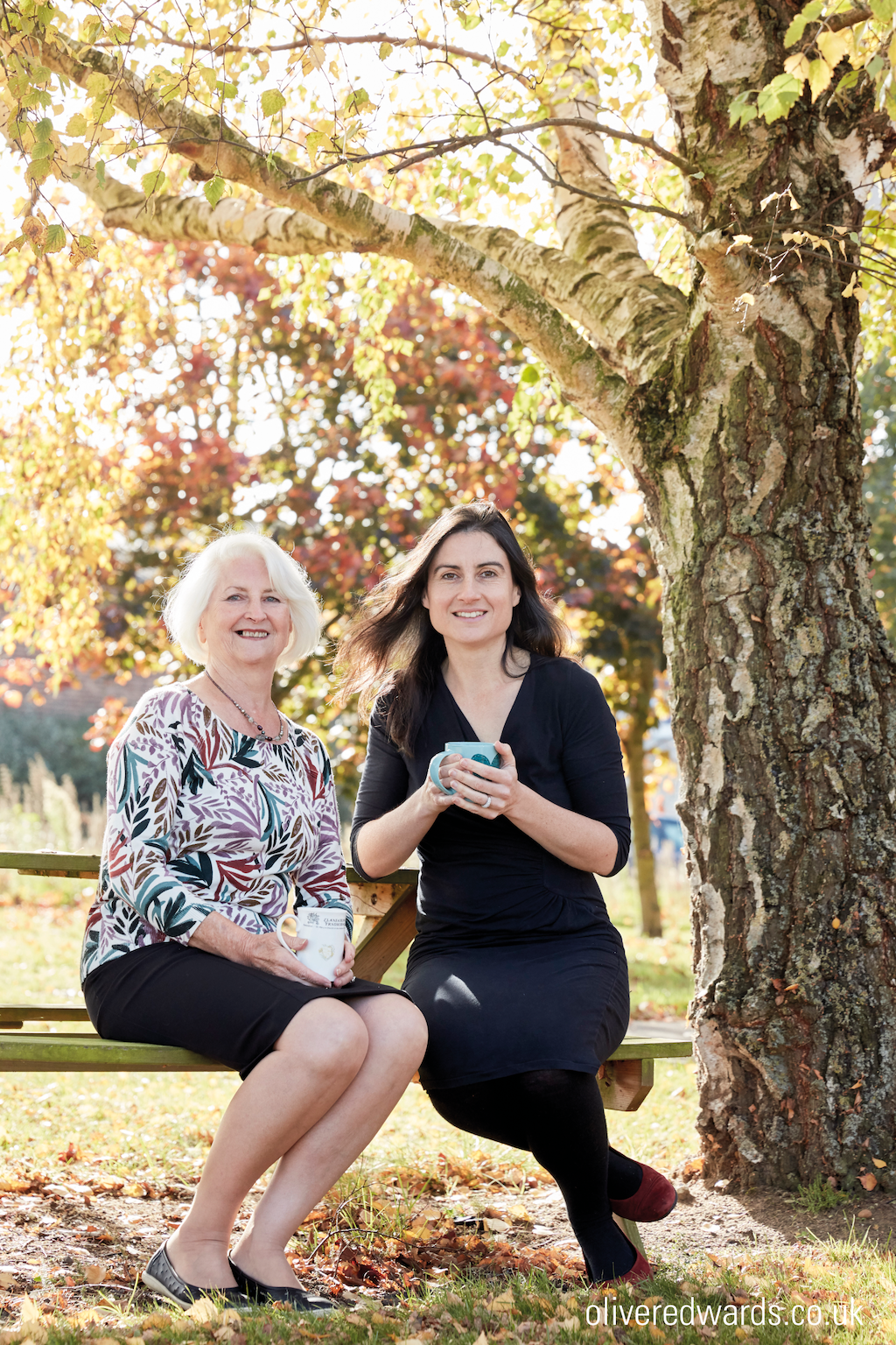 Odylique co-owners Margaret and her daughter Abbie. They are sitting on a bench in the fall under a tree and Abbie is holding a mug.