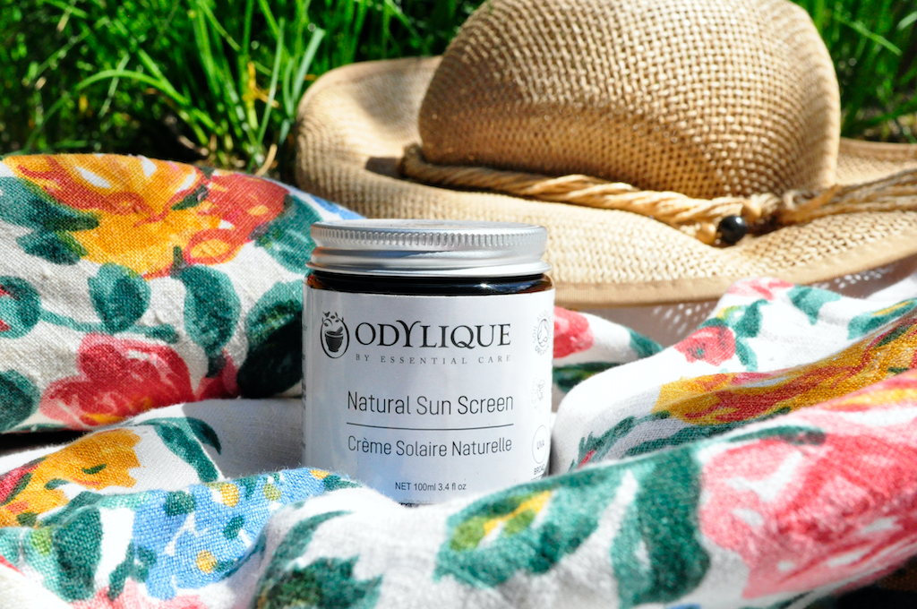 Odylique Natural Mineral Sun Screen SPF 30. Organic Sun Screen. Pictured in an amber glass jar with an aluminium lid, sitting on a floral blanket in the grass with a straw hat behind.