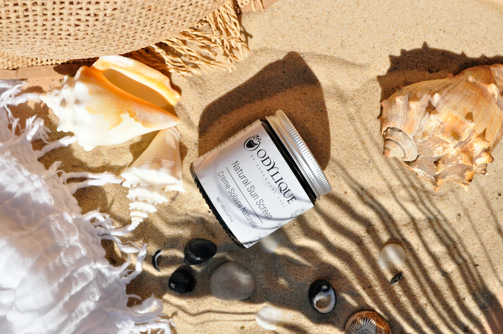 Odylique Natural Mineral Sun Screen SPF 30. Organic Sun Screen. Pictured in an amber glass jar with an aluminium lid, sitting in the sand surrounded by seashells.