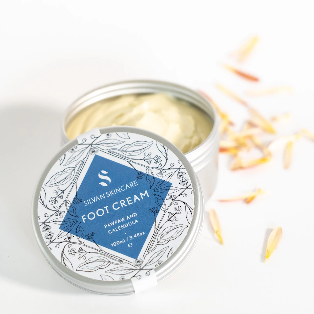 Silvan Skincare Foot Cream. Vegan foot cream. The cream is packaged in an aluminium tub with a delicately illustrated white and blue label. The tub is open with the yellow cream showing and some calendula petals sitting next to it.
