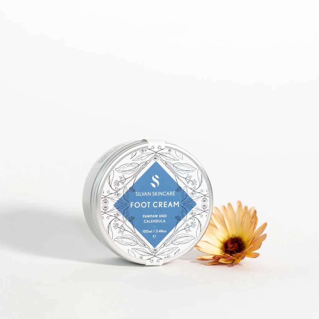 Silvan Skincare Foot Cream. Cruelty free foot cream. The cream is packaged in an aluminium tub with a delicately illustrated white and blue label. A calendula flower is sitting next to the tub.