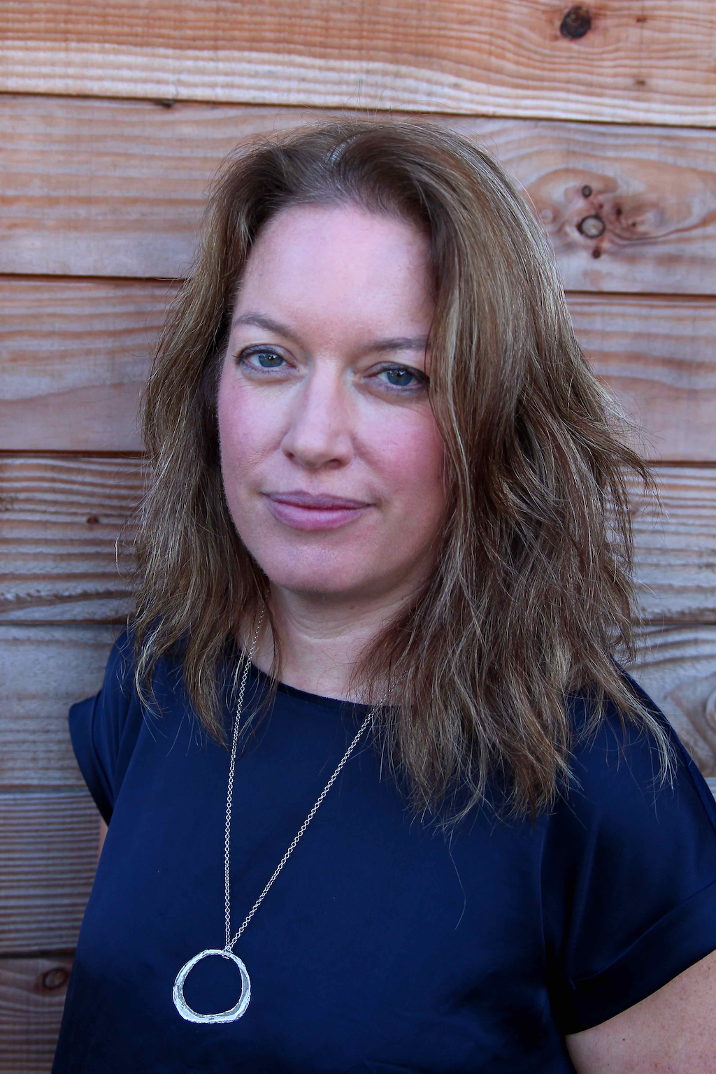 Silvan Skincare Founder Marian. Marian is pictured in a blue shirt with a silver necklace. She has light brown hair with streaks of grey and is standing in front of a wood panelled wall.
