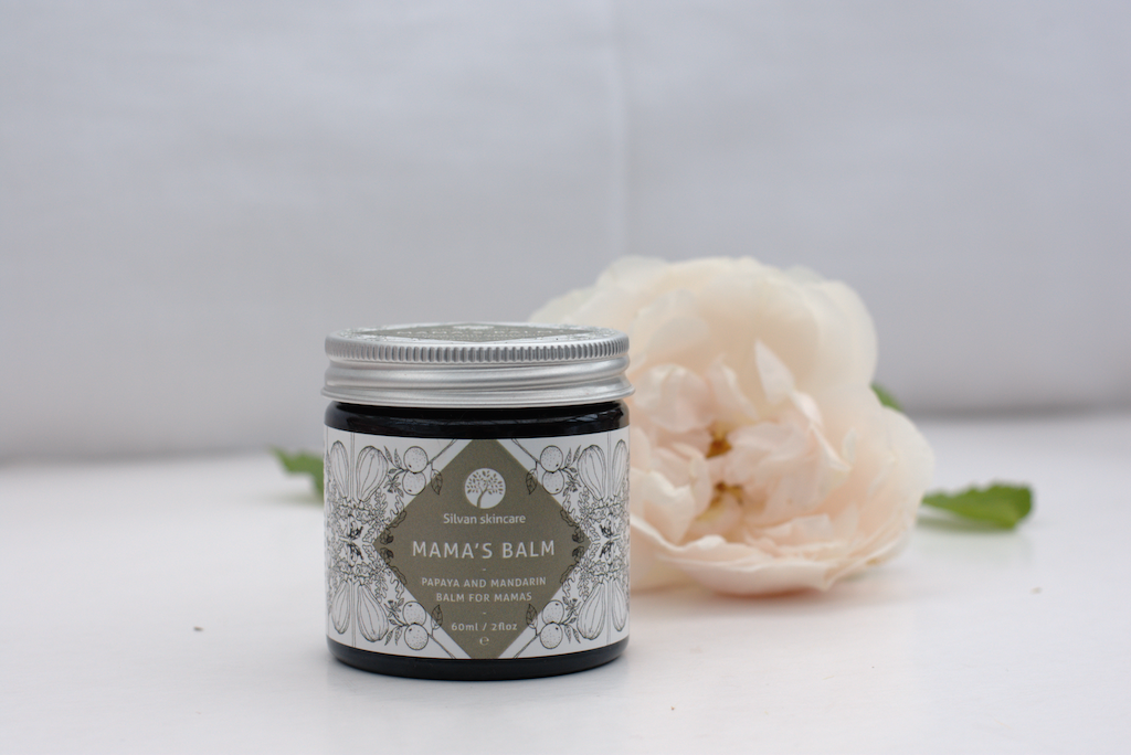 Silvan Skincare Mama's Balm. Plastic free pregnancy products. Pictured in an amber glass jar with an aluminium lid and a delicately illustrated white and grey label, with a white flower sitting behind the jar.