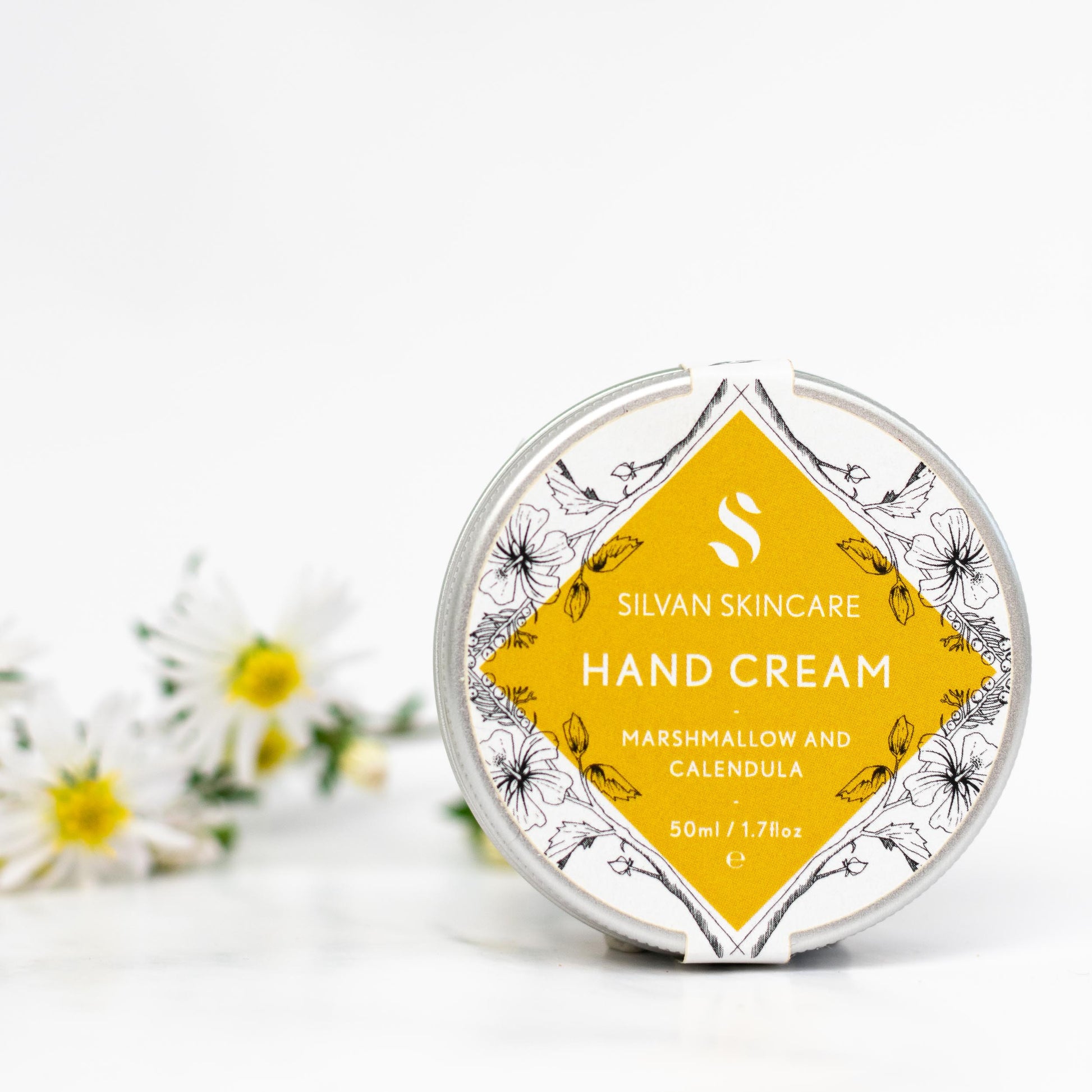 silvan skincare hand cream on a white background with some daisy-like calendula flowers blurred in the background. the hand cream is in an aluminium pot with a white and yellow labels which reads: silvan skincare hand cream marshmallow and calendula 50ml